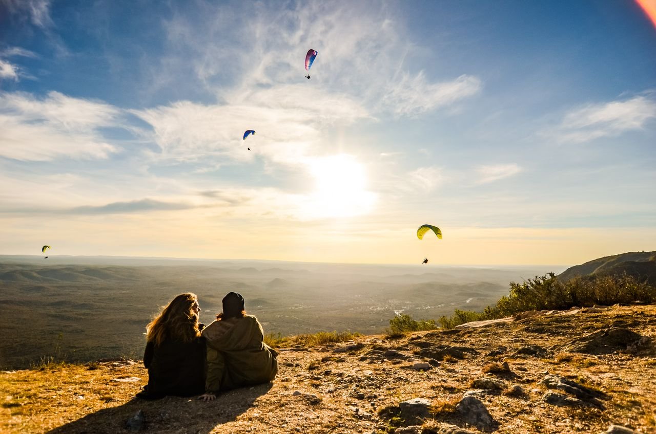 A couple sits on a hill watching paragliders against a stunning sunset backdrop.